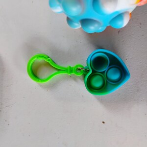 May include: A green and blue heart-shaped fidget toy with three circular indentations. The toy has a green clip attached. A blue and white ball-shaped fidget toy is in the background.