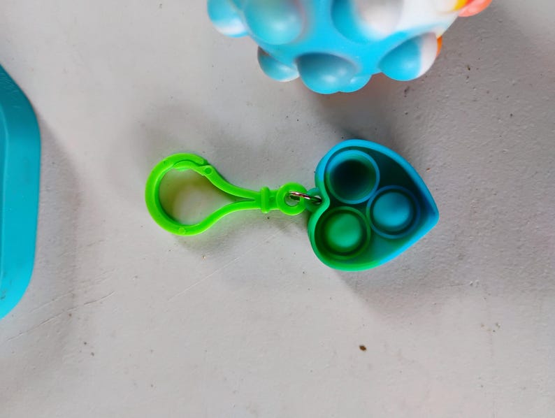 May include: A green and blue heart-shaped fidget toy with three circular indentations. The toy has a green clip attached. A blue and white ball-shaped fidget toy is in the background.