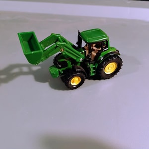 Puede incluir: Un tractor de juguete verde y negro con un cargador frontal. El tractor tiene ruedas amarillas y una cabina detallada. El cargador frontal está levantado y el tractor está sobre un fondo gris claro.