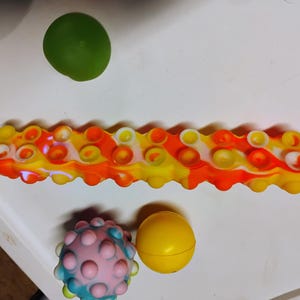 May include: A multi-coloured caterpillar-shaped sensory toy in yellow, orange, and white, with circular bubble-like features. A green sphere, a pink and blue spiky ball, and a yellow sphere are also present.