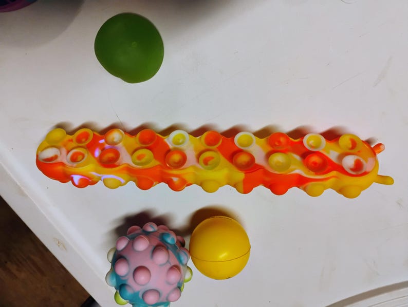 May include: A multi-coloured caterpillar-shaped sensory toy in yellow, orange, and white, with circular bubble-like features. A green sphere, a pink and blue spiky ball, and a yellow sphere are also present.