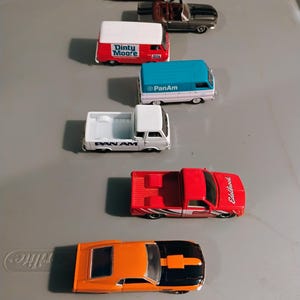 May include: A collection of toy cars in various colors, including red, orange, black, and white. The cars include a red pickup truck with "Edelbrock" text, a red van with "Dinty Moore" text, and a blue and white van with "PanAm" text.