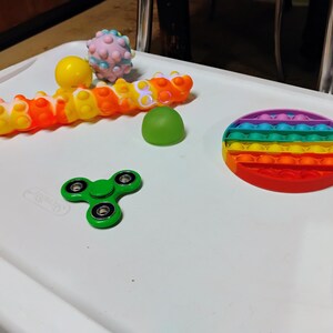 May include: A selection of sensory toys in various colours. The toys include a rainbow pop-it, a green fidget spinner, a caterpillar toy, a yellow ball, a green dome, and a pink spiky ball. They are arranged on a white surface.