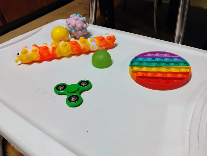 May include: A selection of sensory toys in various colours. The toys include a rainbow pop-it, a green fidget spinner, a caterpillar toy, a yellow ball, a green dome, and a pink spiky ball. They are arranged on a white surface.