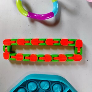 May include: A collection of colorful fidget toys. A green and orange articulated fidget toy is in the center. A rainbow-colored ring fidget toy is at the top. A blue pop-it toy is at the bottom.