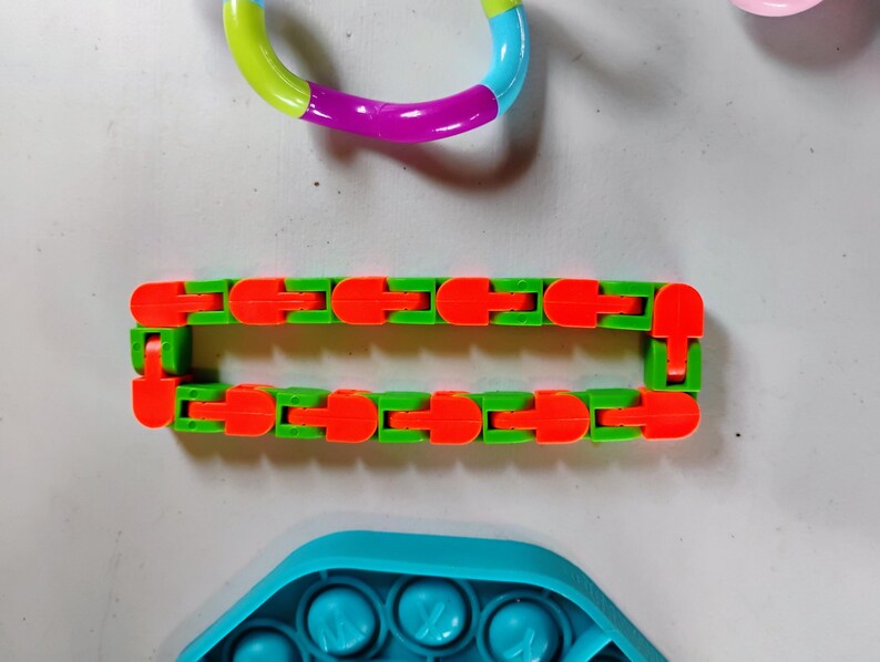 May include: A collection of colorful fidget toys. A green and orange articulated fidget toy is in the center. A rainbow-colored ring fidget toy is at the top. A blue pop-it toy is at the bottom.