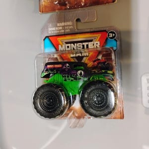 May include: A green and black Grave Digger monster truck toy in a clear plastic package. The toy truck has large black tires and a skull graphic. The packaging includes the text "MONSTER JAM" and a warning about small parts.