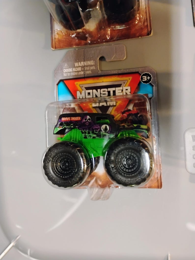 May include: A green and black Grave Digger monster truck toy in a clear plastic package. The toy truck has large black tires and a skull graphic. The packaging includes the text "MONSTER JAM" and a warning about small parts.