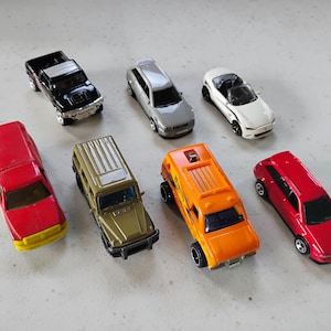 Könnte beinhalten: Eine Sammlung von sieben Spielzeugautos aus Metall, bestehend aus einem schwarzen Pickup, einer silbernen Limousine, einem weißen Cabriolet, einem roten LKW mit gelber Stoßstange, einem beigen SUV, einem orangefarbenen Van und einem roten Schrägheckmodell.