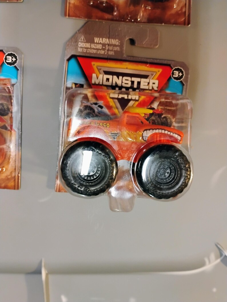 May include: Orange and yellow Monster Jam truck toy in a clear plastic package. The truck has large black tires and the name "Pro Loco" printed on the side. The package has the Monster Jam logo and a warning about small parts.