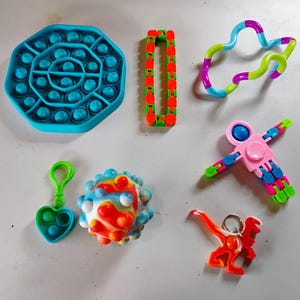 May include: A collection of colorful fidget toys, including a blue octagon-shaped pop-it, a red and green chain, a multi-colored twisty toy, a pink and blue spinner, a heart-shaped pop-it keychain, a spiky ball, and an orange dinosaur keychain.