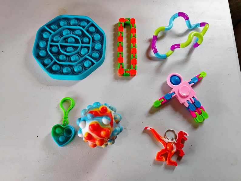 May include: A collection of colorful fidget toys, including a blue octagon-shaped pop-it, a red and green chain, a multi-colored twisty toy, a pink and blue spinner, a heart-shaped pop-it keychain, a spiky ball, and an orange dinosaur keychain.