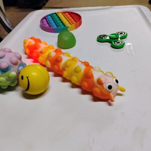 May include: A selection of sensory toys, including a rainbow pop-it, a green fidget spinner, a yellow smiley face ball, a spiky ball, and a caterpillar-shaped toy with orange, yellow, and white segments.