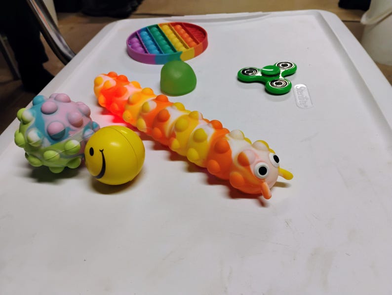 May include: A selection of sensory toys, including a rainbow pop-it, a green fidget spinner, a yellow smiley face ball, a spiky ball, and a caterpillar-shaped toy with orange, yellow, and white segments.