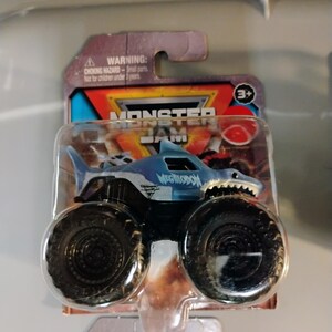 May include: A blue and white Megalodon monster truck toy with large black tires. The truck is part of the Monster Jam series and is packaged in a clear plastic blister pack. The packaging includes a warning about small parts and is not for children under 3 years.