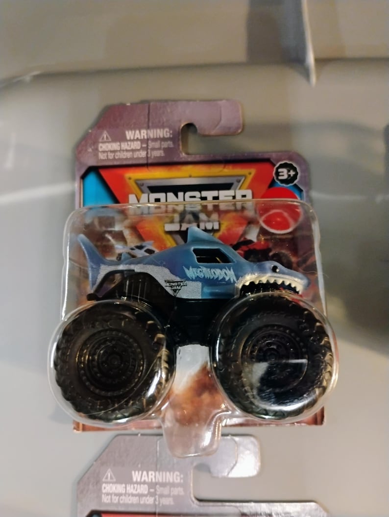 May include: A blue and white Megalodon monster truck toy with large black tires. The truck is part of the Monster Jam series and is packaged in a clear plastic blister pack. The packaging includes a warning about small parts and is not for children under 3 years.