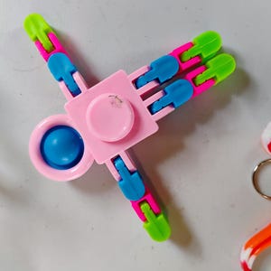 May include: A pink, blue, and green fidget spinner toy. The spinner has a central pink square with a pink circle in the middle and a blue circle in the center. The arms are pink, blue, and green.