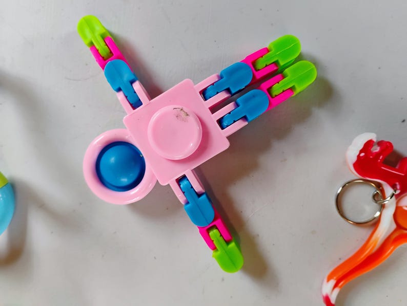 May include: A pink, blue, and green fidget spinner toy. The spinner has a central pink square with a pink circle in the middle and a blue circle in the center. The arms are pink, blue, and green.