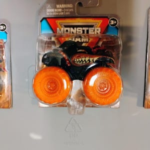 May include: A black Monster Jam truck toy with orange wheels is displayed in its original packaging. The truck features a menacing design with a toothy grin and the Monster Jam logo. The packaging includes a warning about small parts and is suitable for ages 3 and up.