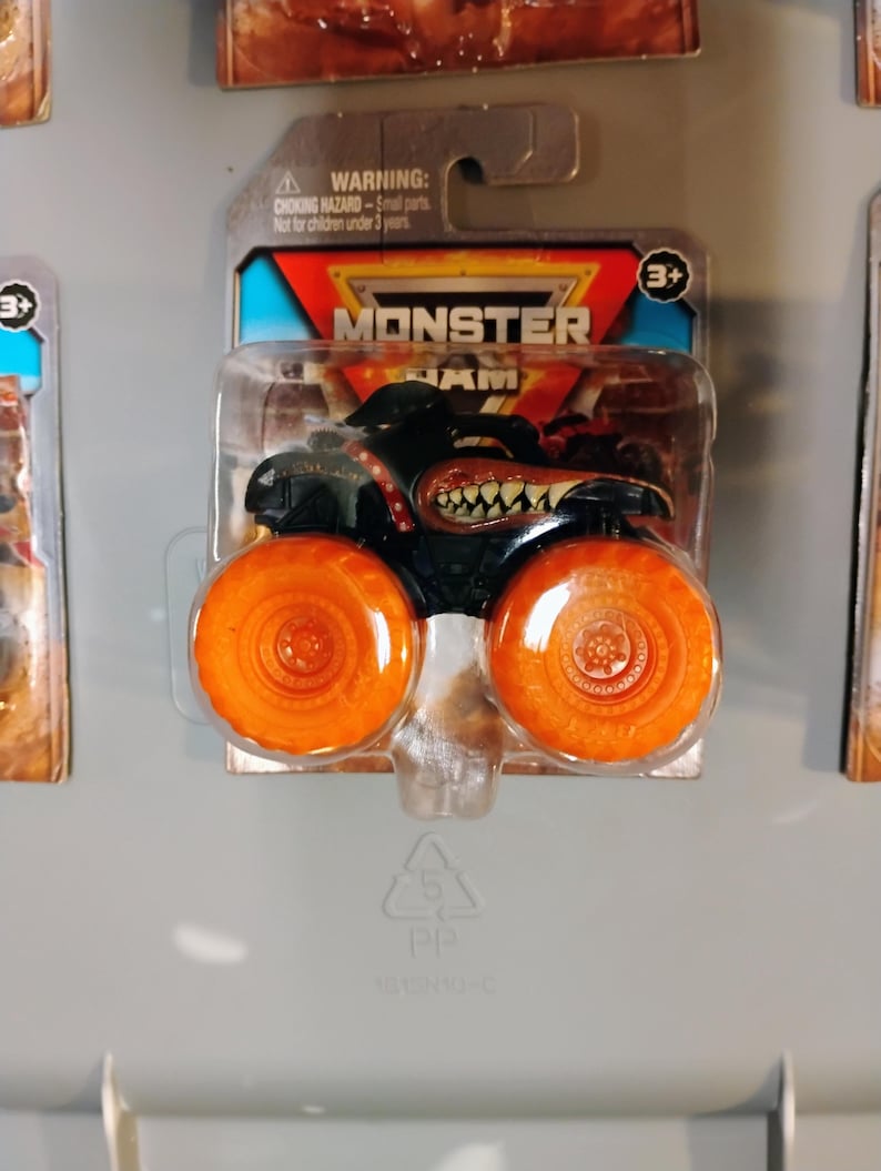 May include: A black Monster Jam truck toy with orange wheels is displayed in its original packaging. The truck features a menacing design with a toothy grin and the Monster Jam logo. The packaging includes a warning about small parts and is suitable for ages 3 and up.