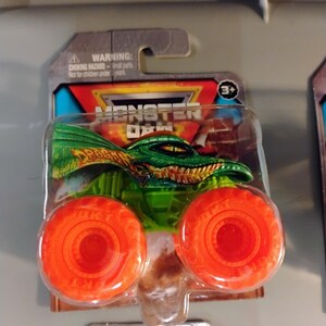May include: A Monster Jam toy truck featuring a green and gold dragon design. The truck has large orange wheels and is packaged in a clear plastic blister pack. The packaging includes the text "WARNING: CHOKING HAZARD - Small parts. Not for children under 3 years."