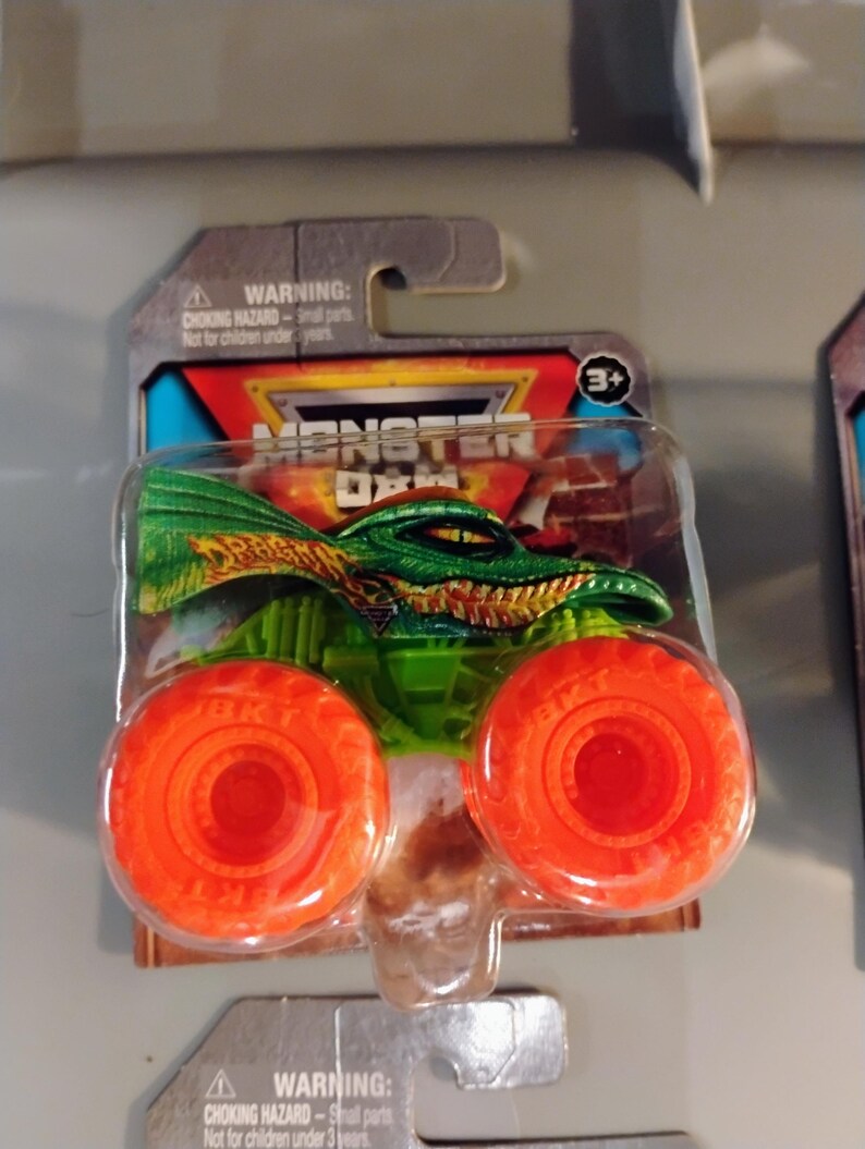 May include: A Monster Jam toy truck featuring a green and gold dragon design. The truck has large orange wheels and is packaged in a clear plastic blister pack. The packaging includes the text "WARNING: CHOKING HAZARD - Small parts. Not for children under 3 years."