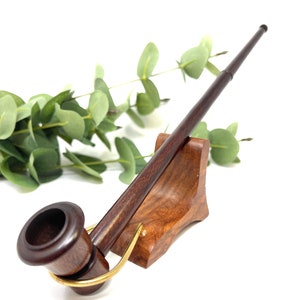 Könnte beinhalten: Eine dunkelbraune Holzpfeife mit einem Messingband und einem Holzständer. Die Pfeife liegt auf dem Ständer, die Schale zeigt zur Kamera.