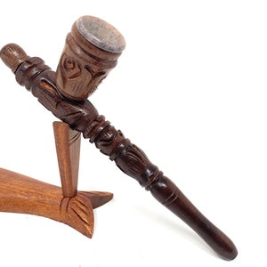 May include: A wooden smoking pipe with intricate carvings, featuring a bowl with a dark brown finish and a long, slender stem. The pipe is displayed on a wooden stand shaped like a fish.