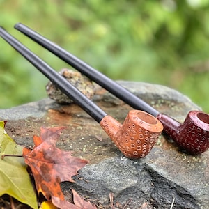 Peut inclure: Deux pipes à fumer en bois avec des tiges noires. L'une est brune avec un motif circulaire et l'autre est rouge foncé avec un motif similaire.