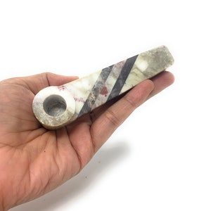 Puede incluir: Una mano sosteniendo una pipa de mármol blanco con rayas negras y rojas. La pipa mide aproximadamente 13 cm de largo y tiene una cazoleta redonda.