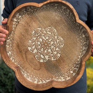 K&ouml;nnte beinhalten: Ein gro&szlig;es, h&ouml;lzernes Serviertablett mit einem gewellten Rand und aufwendig geschnitzten Blumenmustern. Das Tablett ist aus hellbraunem Holz gefertigt und hat eine wei&szlig;e Lasur.