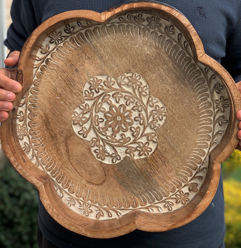 K&ouml;nnte beinhalten: Ein gro&szlig;es, h&ouml;lzernes Serviertablett mit einem gewellten Rand und aufwendig geschnitzten Blumenmustern. Das Tablett ist aus hellbraunem Holz gefertigt und hat eine wei&szlig;e Lasur.