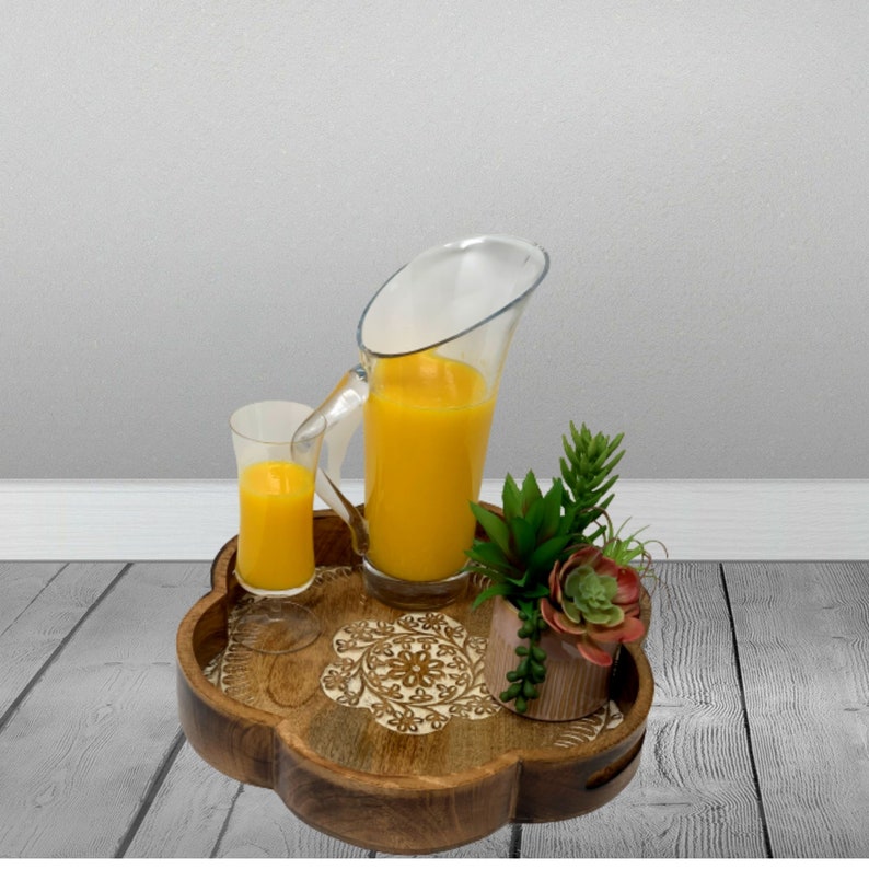 K&ouml;nnte beinhalten: Ein Holztablett mit einem geschnitzten Design, auf dem ein Glaskrug und ein Glas Orangensaft stehen. Eine kleine Topfpflanze mit Sukkulenten befindet sich ebenfalls auf dem Tablett.