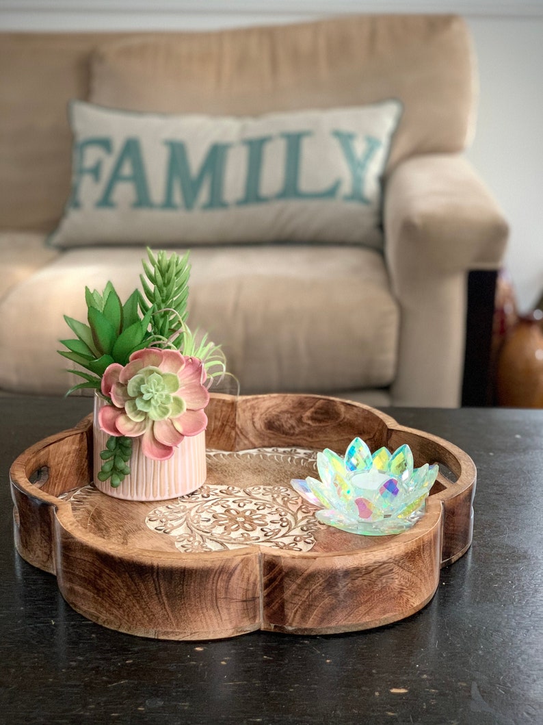 K&ouml;nnte beinhalten: Ein Holztablett mit einem geschnitzten Design zeigt einen rosafarbenen Sukkulententopf und einen regenbogenfarbenen Glas-Lotus-Kerzenhalter. Das Tablett befindet sich auf einer dunklen Oberfl&auml;che.