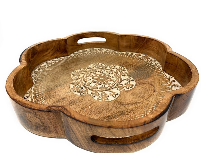 K&ouml;nnte beinhalten: Ein rundes Serviertablett aus Holz mit einem geschnitzten Blumenmuster. Das Tablett hat zwei Griffe an den Seiten und ist aus hellbraunem Holz gefertigt.
