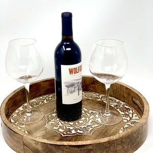 K&ouml;nnte beinhalten: Ein rundes Serviertablett aus Holz mit aufwendigen floralen Schnitzereien in hellbrauner Farbe. Auf dem Tablett stehen zwei Weingl&auml;ser und eine Flasche Rotwein. Das Flaschenetikett lautet "Wolfson Cellars Red Wine California".