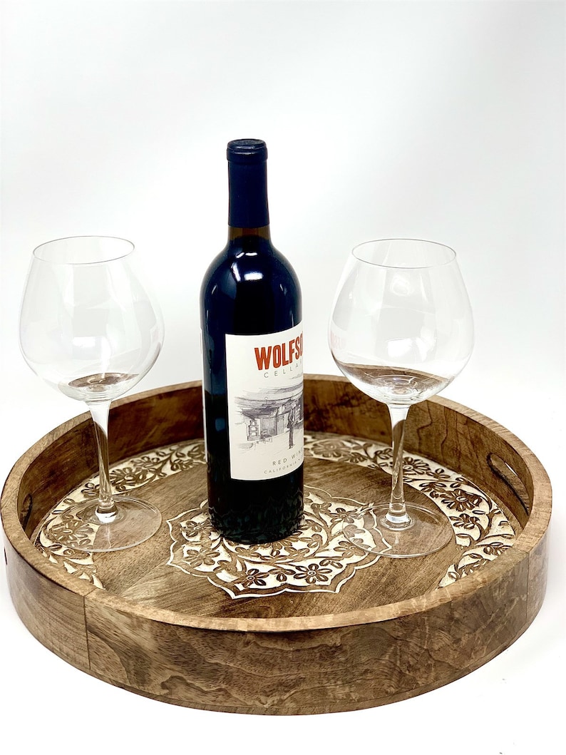 K&ouml;nnte beinhalten: Ein rundes Serviertablett aus Holz mit aufwendigen floralen Schnitzereien in hellbrauner Farbe. Auf dem Tablett stehen zwei Weingl&auml;ser und eine Flasche Rotwein. Das Flaschenetikett lautet "Wolfson Cellars Red Wine California".