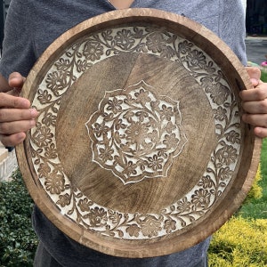 K&ouml;nnte beinhalten: Ein rundes Holztablett mit einem geschnitzten Blumenmuster. Das Tablett ist braun mit wei&szlig;en Akzenten und hat einen erh&ouml;hten Rand.