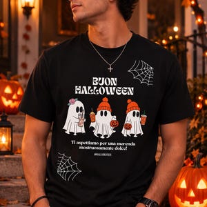 Può includere: Una t-shirt nera con la scritta BUON HALLOWEEN e tre illustrazioni di fantasmi bianchi che indossano cappelli e tengono in mano bevande e zucche. Sotto i fantasmi c'è il testo Ti aspettiamo per una merenda mostruosamente dolce! e @REALLYCREATISTE, con grafiche di ragnatele bianche.