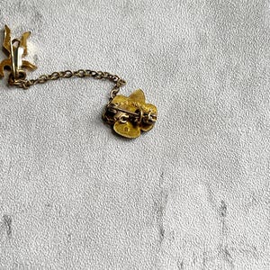 Puede incluir: Broche dorado vintage con dise&ntilde;o floral, conectado por una delicada cadena a un cierre decorativo. El broche tiene un pasador en la parte posterior para una fijaci&oacute;n segura. El metal tiene un aspecto ligeramente envejecido.