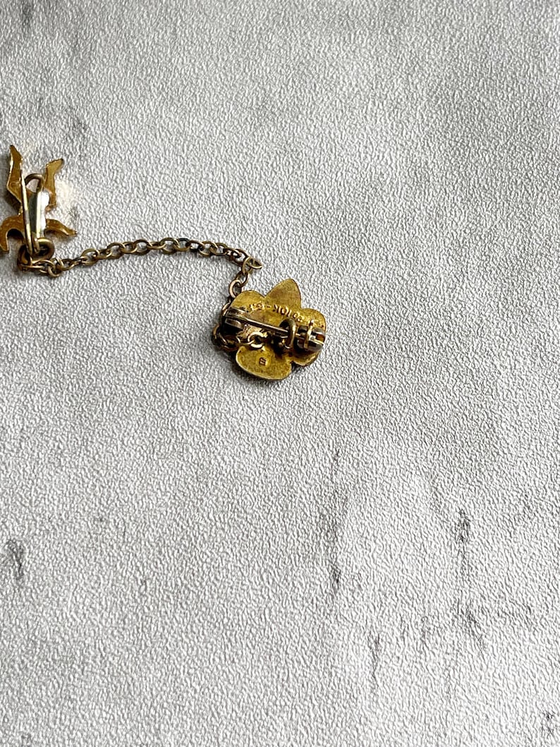 Puede incluir: Broche dorado vintage con dise&ntilde;o floral, conectado por una delicada cadena a un cierre decorativo. El broche tiene un pasador en la parte posterior para una fijaci&oacute;n segura. El metal tiene un aspecto ligeramente envejecido.