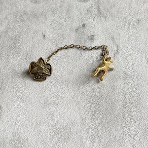 Puede incluir: Un broche vintage dorado con una cadena. Un extremo presenta un dise&ntilde;o de flor de lis, y el otro un peque&ntilde;o caballo corriendo. El broche probablemente est&aacute; hecho de lat&oacute;n o un metal similar. La cadena conecta los dos elementos decorativos.
