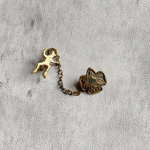 Puede incluir: Conjunto de broches dorados vintage. Una peque&ntilde;a figura est&aacute; conectada por una cadena a un broche en forma de flor. El broche de la flor tiene un dise&ntilde;o detallado con un escudo. El fondo es una superficie texturizada de color gris claro.