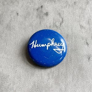 May include: A round, blue button with the name "Humphrey" in white cursive script. The button has a glossy finish and is set against a textured, gray background. The design is simple and the text is the focal point.