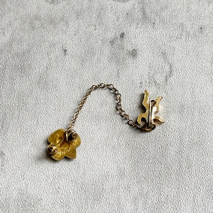 Puede incluir: Un broche vintage dorado con un alfiler en forma de flor y una letra estilizada. El broche est&aacute; conectado por una delicada cadena a un segundo alfiler con un dise&ntilde;o decorativo. El fondo es una superficie texturizada de color gris claro.