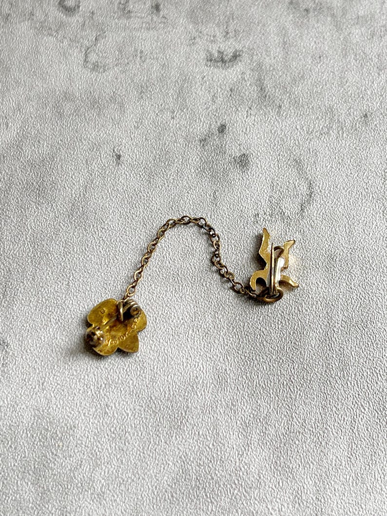 Puede incluir: Un broche vintage dorado con un alfiler en forma de flor y una letra estilizada. El broche est&aacute; conectado por una delicada cadena a un segundo alfiler con un dise&ntilde;o decorativo. El fondo es una superficie texturizada de color gris claro.