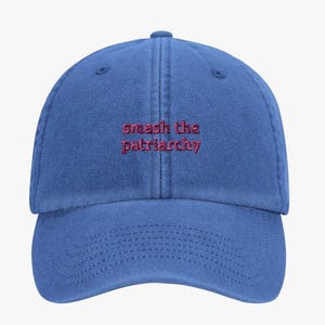May include: A blue baseball cap with a six-panel construction and a curved brim. The front features the embroidered text "smash the patriarchy" in a dark red thread, centred above the brim. The fabric has a slightly weathered, matte texture.