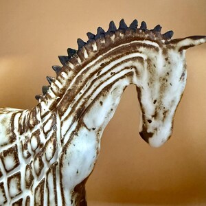 May include: A ceramic zebra figurine with a cream-colored body and brown stripes. The zebra has a detailed mane and a curved neck, showcasing a handcrafted aesthetic. The figurine is a decorative item.