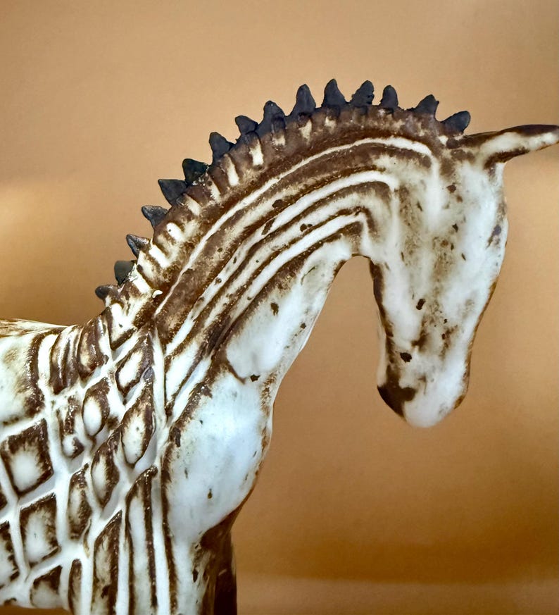 May include: A ceramic zebra figurine with a cream-colored body and brown stripes. The zebra has a detailed mane and a curved neck, showcasing a handcrafted aesthetic. The figurine is a decorative item.