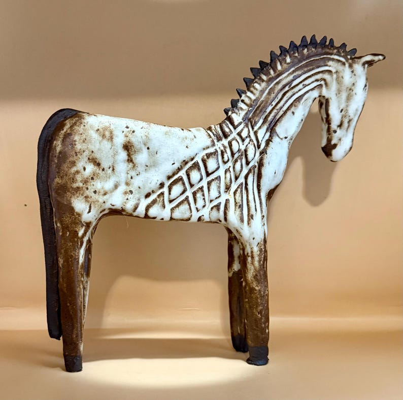 May include: A ceramic horse figurine in a standing pose. The horse is primarily white with brown accents, including the mane, tail, and legs. The body features a diamond pattern. The sculpture has a rustic, handcrafted appearance.